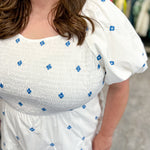 White dress with flowy bottom and smocked top. Round neckline. Short sleeve style. Blue floral embroidered detail. Hits right below the knee.