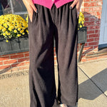 Person wearing a pink cardigan and black pants standing outdoors with a brick wall and yellow flowers in the background.