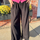 Person wearing a pink cardigan and black pants standing outdoors with a brick wall and yellow flowers in the background.