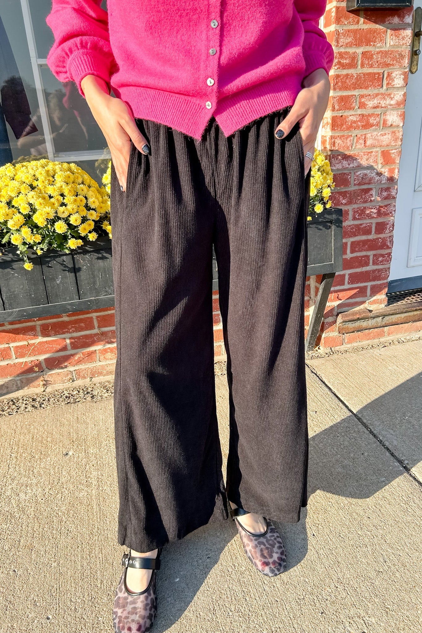 Person wearing a pink cardigan and black pants standing outdoors with a brick wall and yellow flowers in the background.
