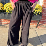 Person wearing a bright pink sweater and dark wide-leg pants standing on a sidewalk with yellow flowers and a brick wall in the background.