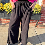 Person wearing a bright pink sweater and dark wide-leg pants standing on a sidewalk with yellow flowers and a brick wall in the background.