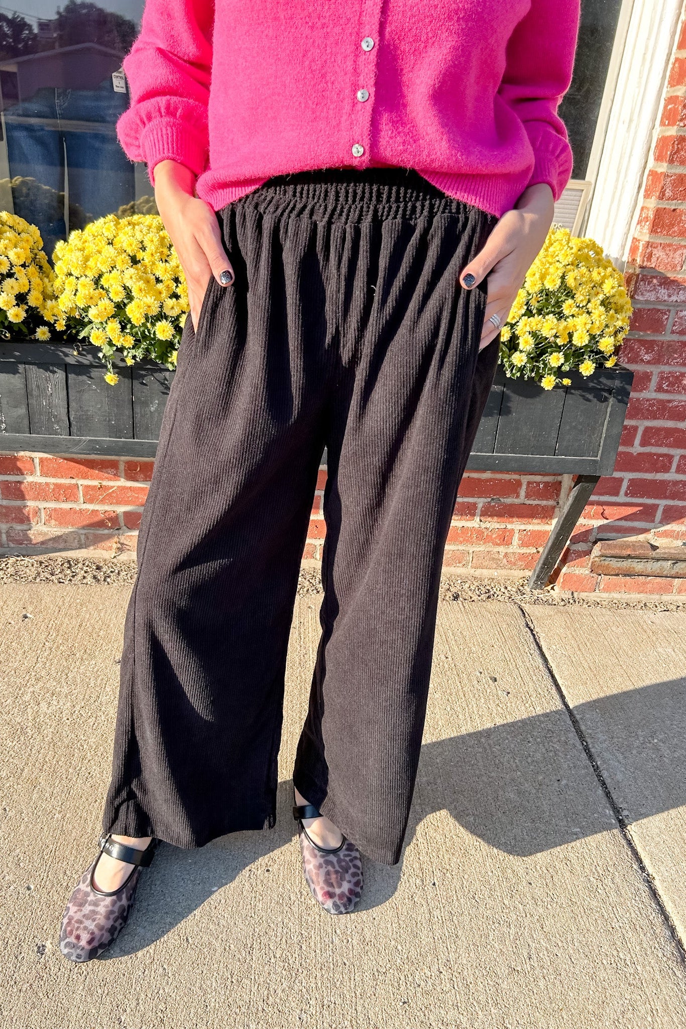 Person wearing a bright pink sweater and dark wide-leg pants standing on a sidewalk with yellow flowers and a brick wall in the background.