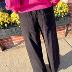 Person wearing a bright pink sweater and black pants standing outdoors with a brick wall and yellow flowers in the background.