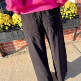 Person wearing a bright pink sweater and black pants standing outdoors with a brick wall and yellow flowers in the background.