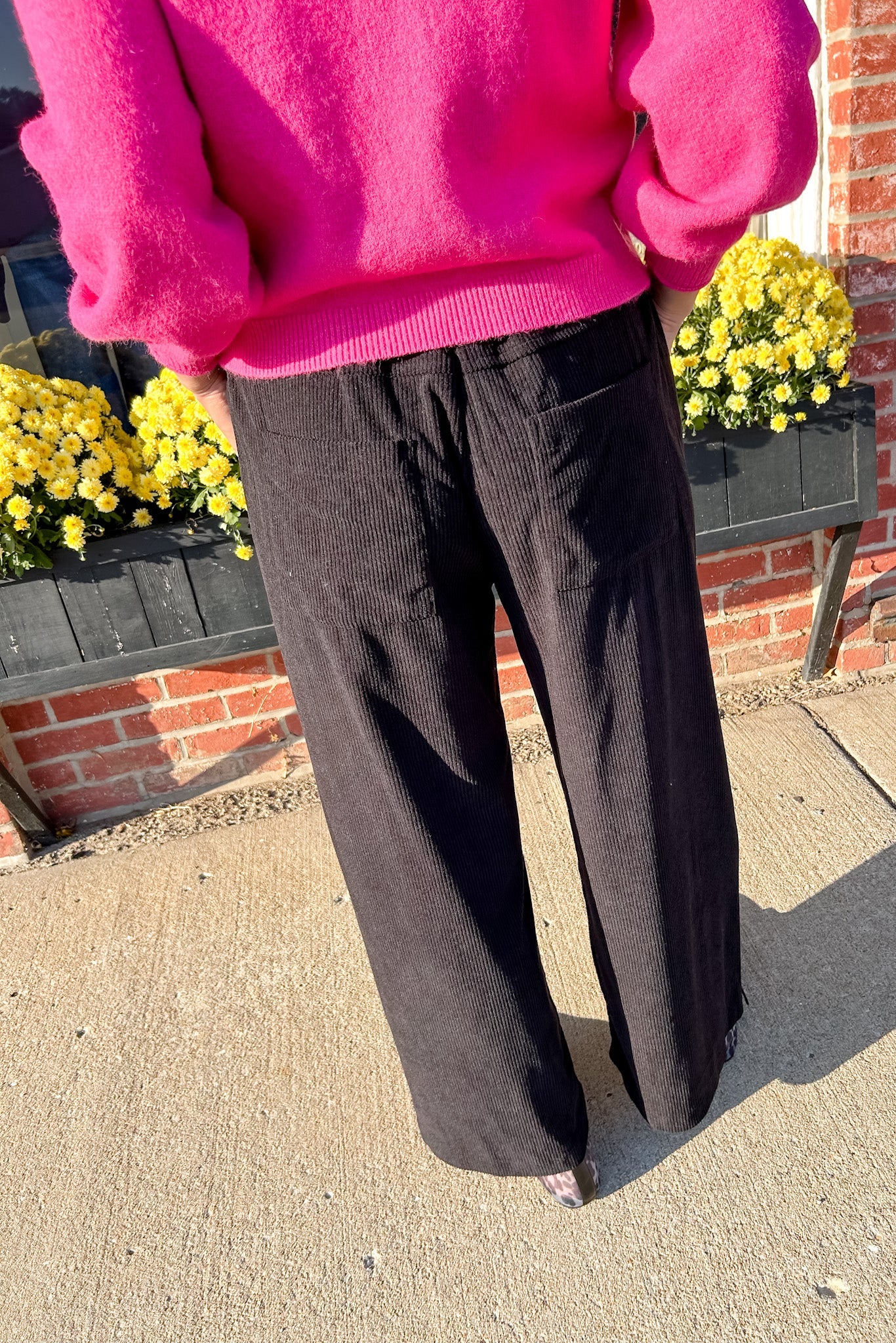 Person wearing a bright pink sweater and black pants standing outdoors with a brick wall and yellow flowers in the background.
