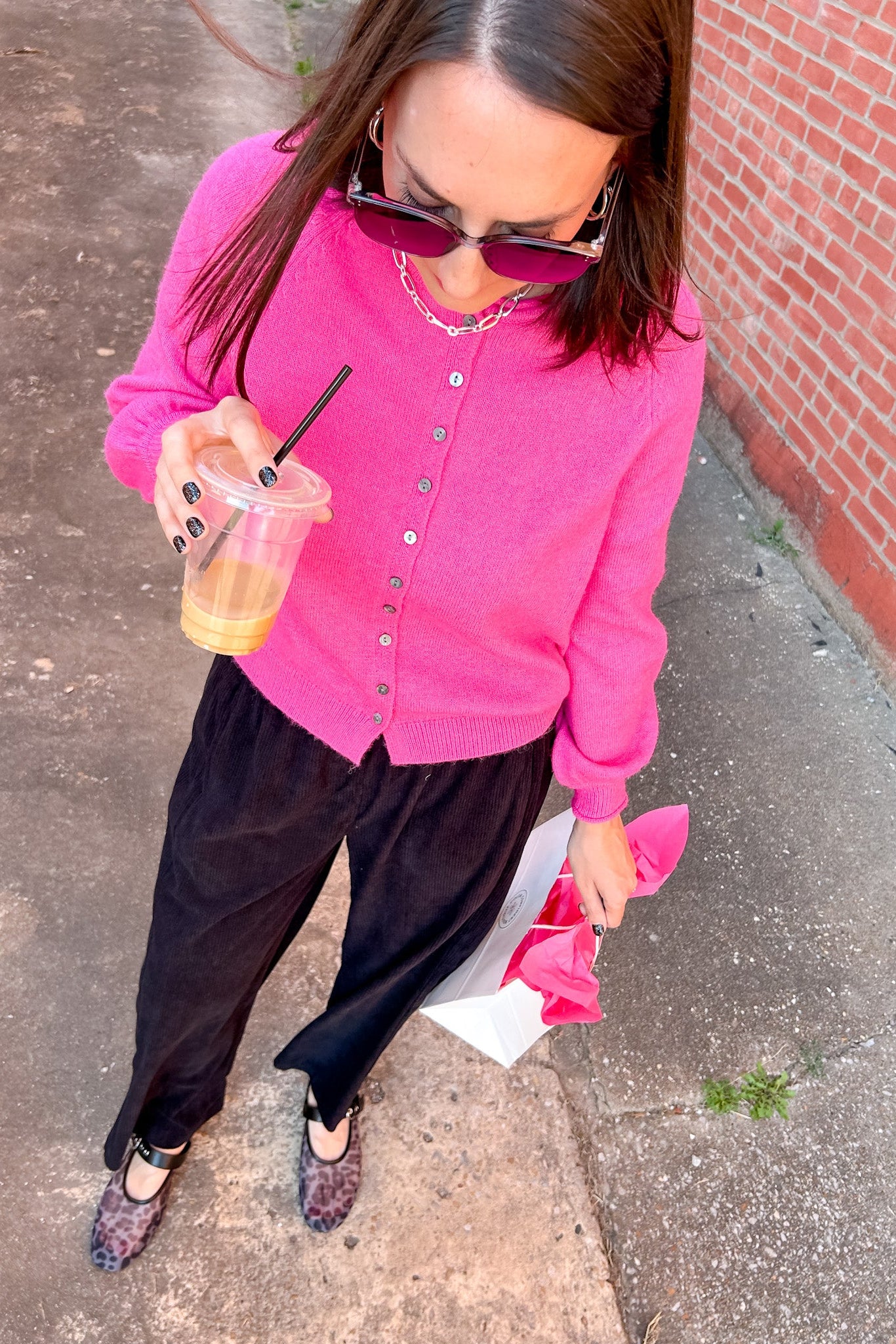 Woman in a pink cardigan and black pants holding a drink on a sidewalk.