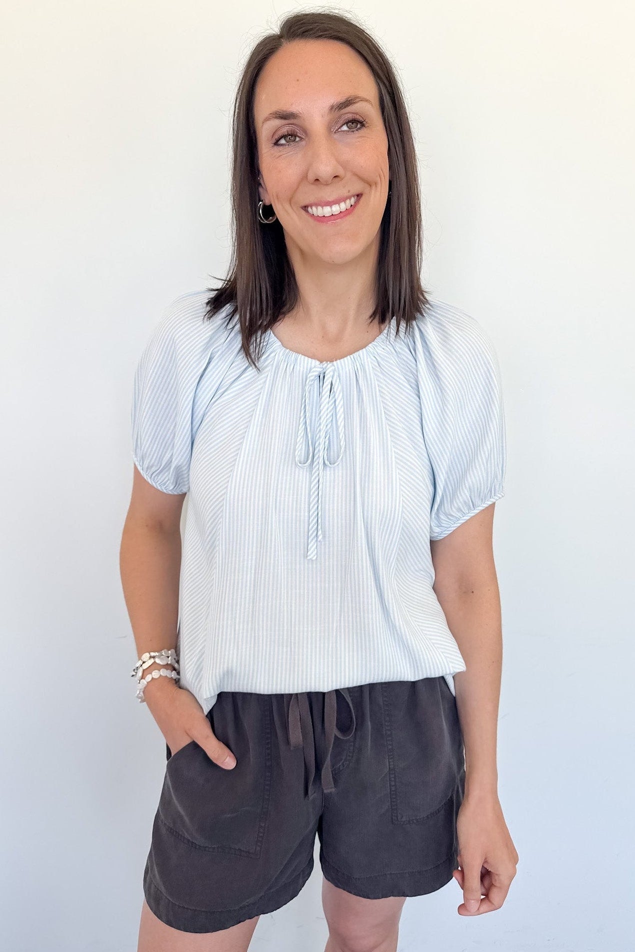 Woman in a light blue striped top that is short sleeve with charcoal grey shorts.