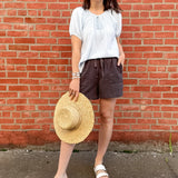 Woman standing in front of a brick wall with a light blue striped top on and grey shorts.