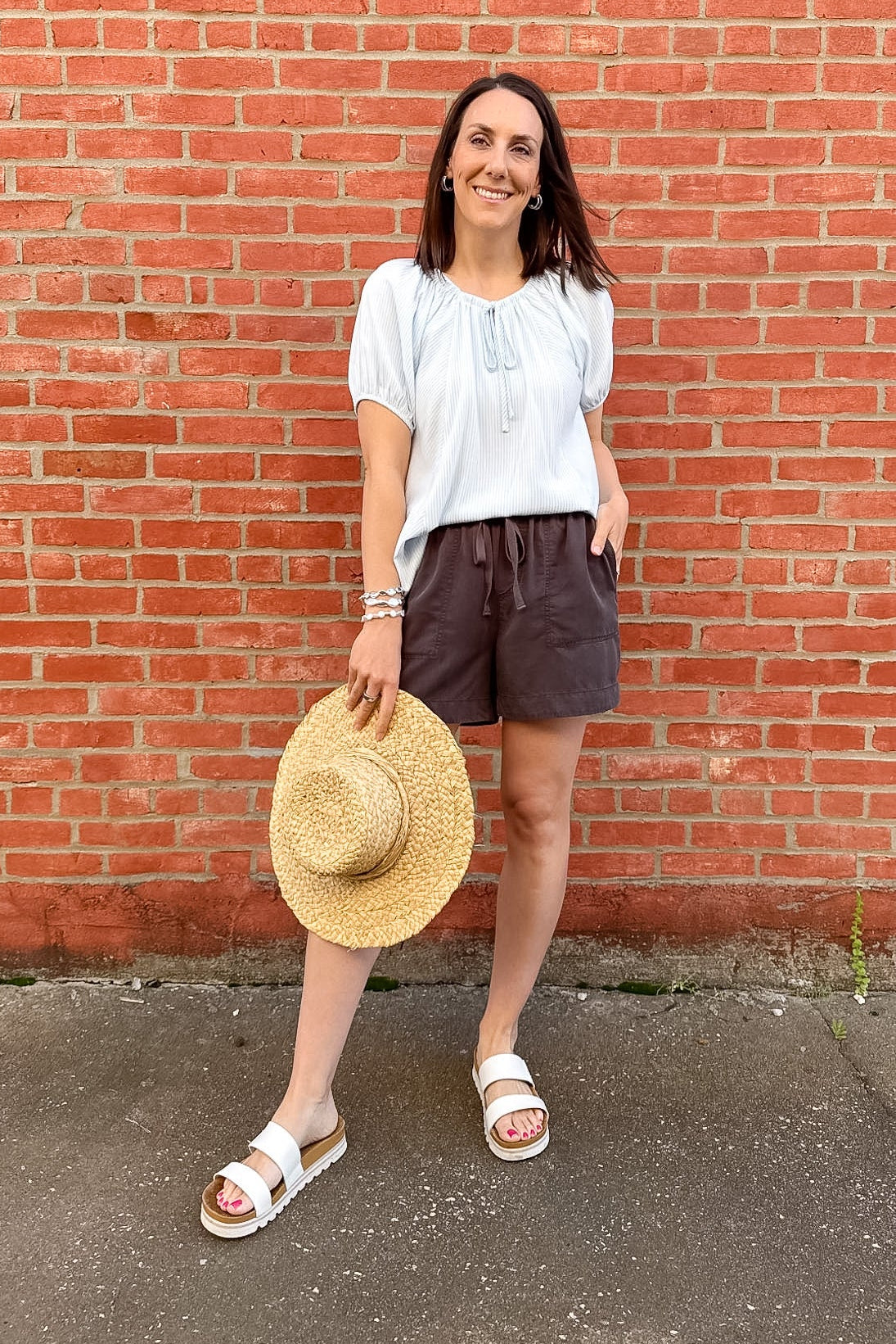Woman standing in front of a brick wall with a light blue striped top on and grey shorts.