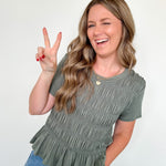 Woman wearing a green ruffled top and blue jeans, making a peace sign.