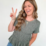 Woman wearing a green ruffled top and blue jeans, making a peace sign.