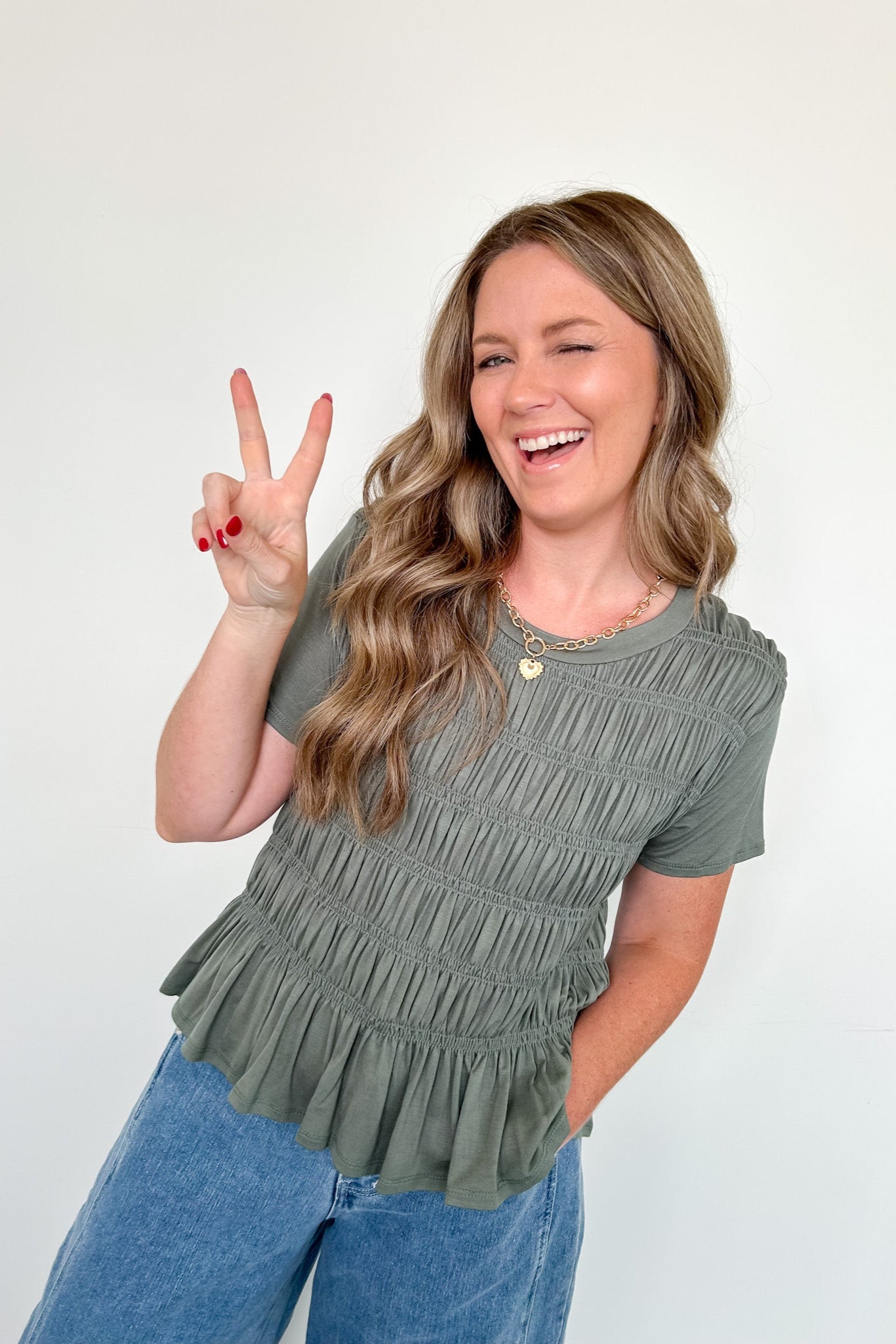 Woman wearing a green ruffled top and blue jeans, making a peace sign.