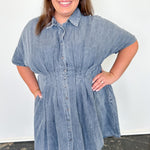 Woman wearing a denim dress standing against a white wall.
