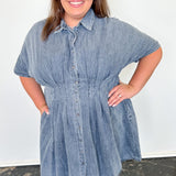 Woman wearing a denim dress standing against a white wall.