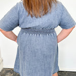Person wearing a blue denim dress standing on a dark rug with a white wall background