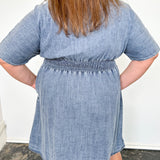 Person wearing a blue denim dress standing on a dark rug with a white wall background