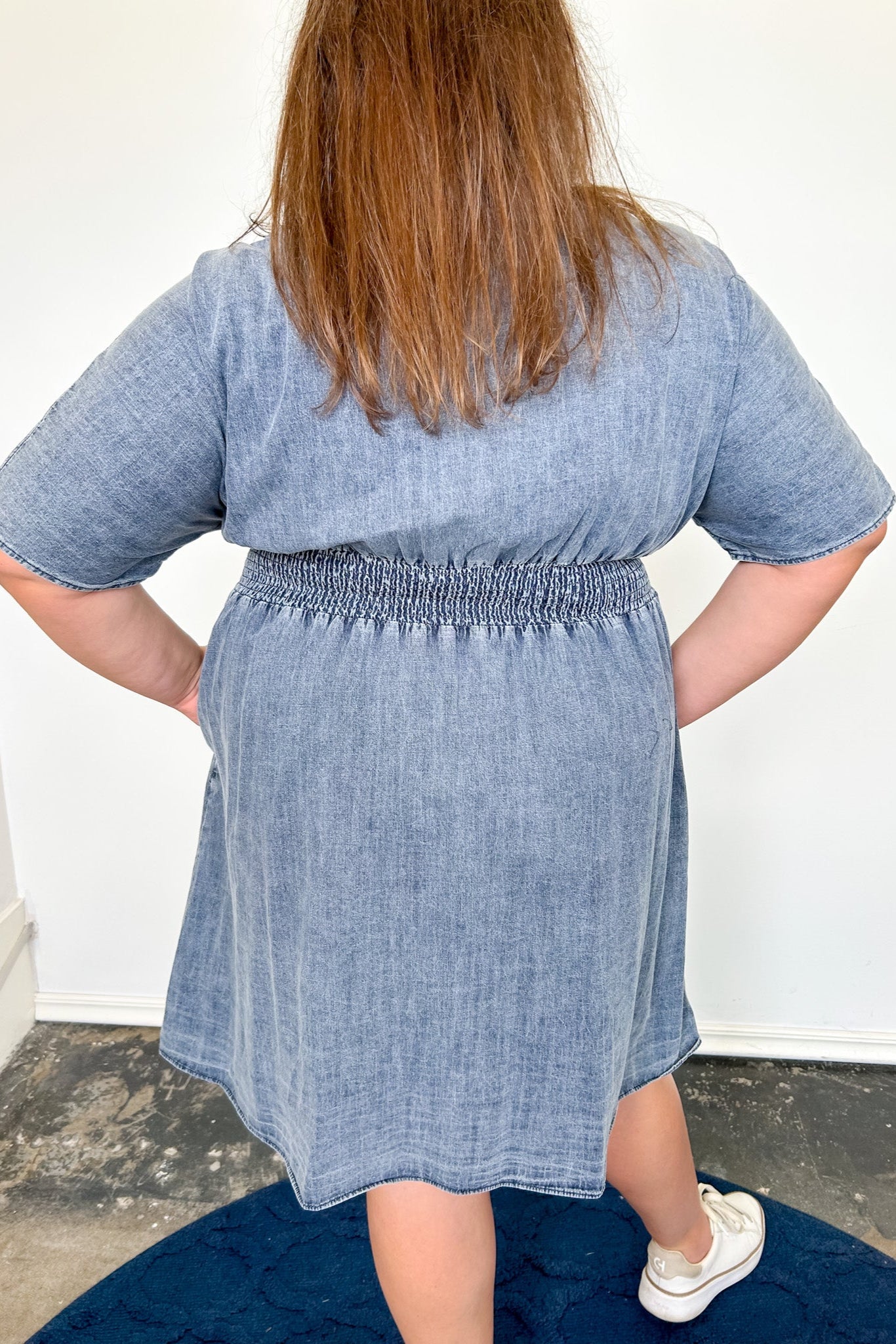 Person wearing a blue denim dress standing on a dark rug with a white wall background