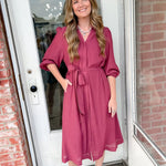 Woman in a pink dress standing in front of a door with brick walls.