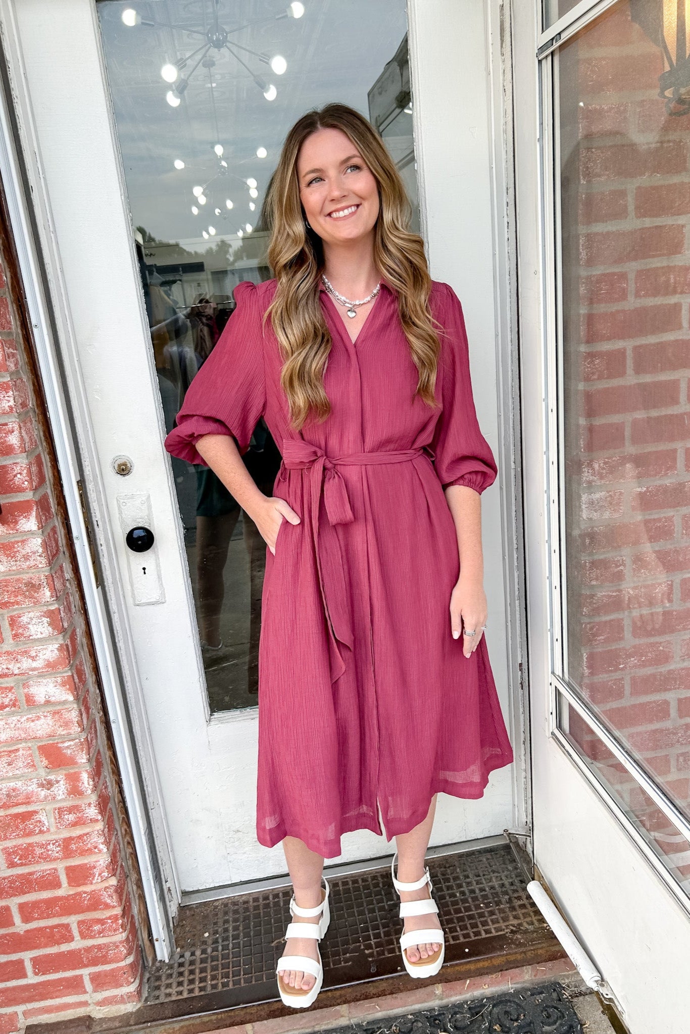 Woman in a pink dress standing in front of a door with brick walls.