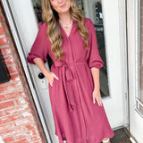 Woman in a pink dress standing in front of a door with brick walls.