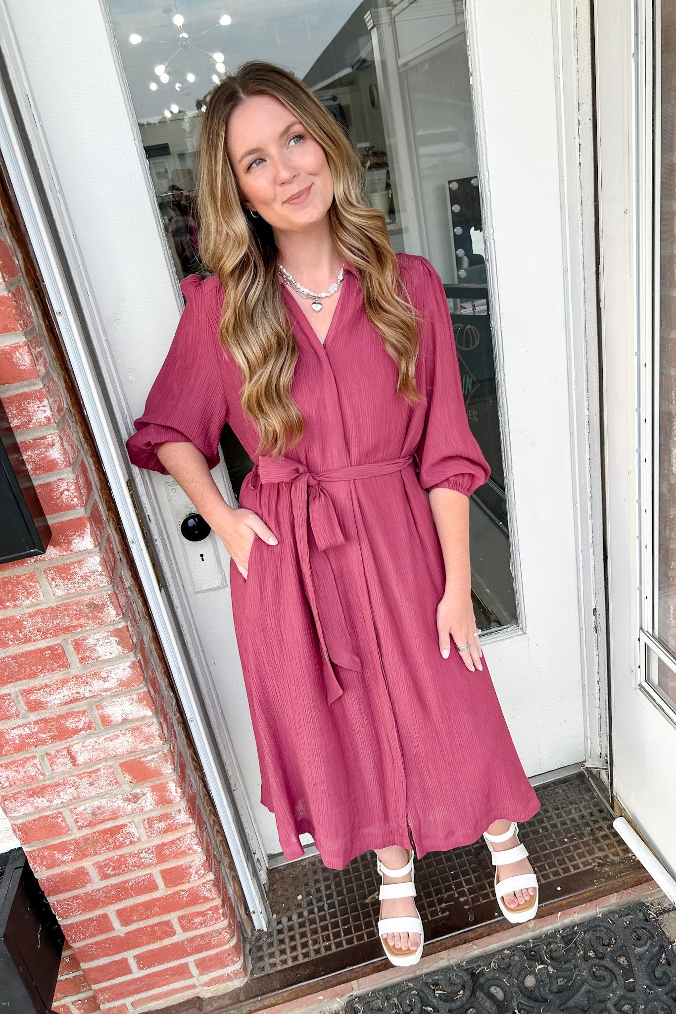 Woman in a pink dress standing in front of a door with brick walls.