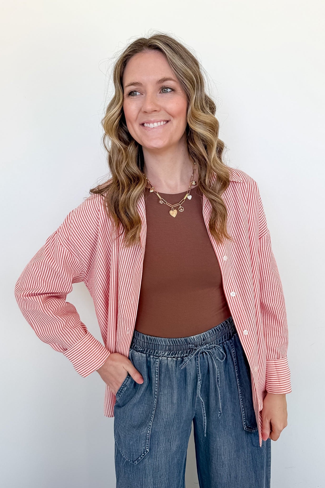 Woman wearing a pink striped shirt, brown top, and blue jeans against a white background