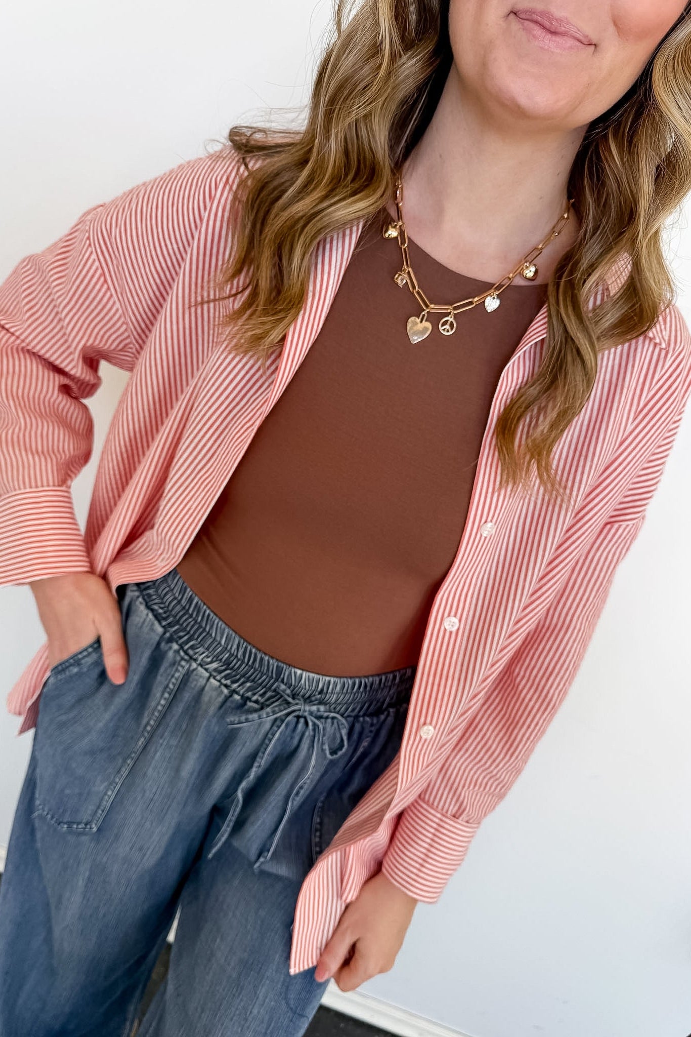 Woman wearing a pink striped shirt over a brown top and blue jeans against a white background