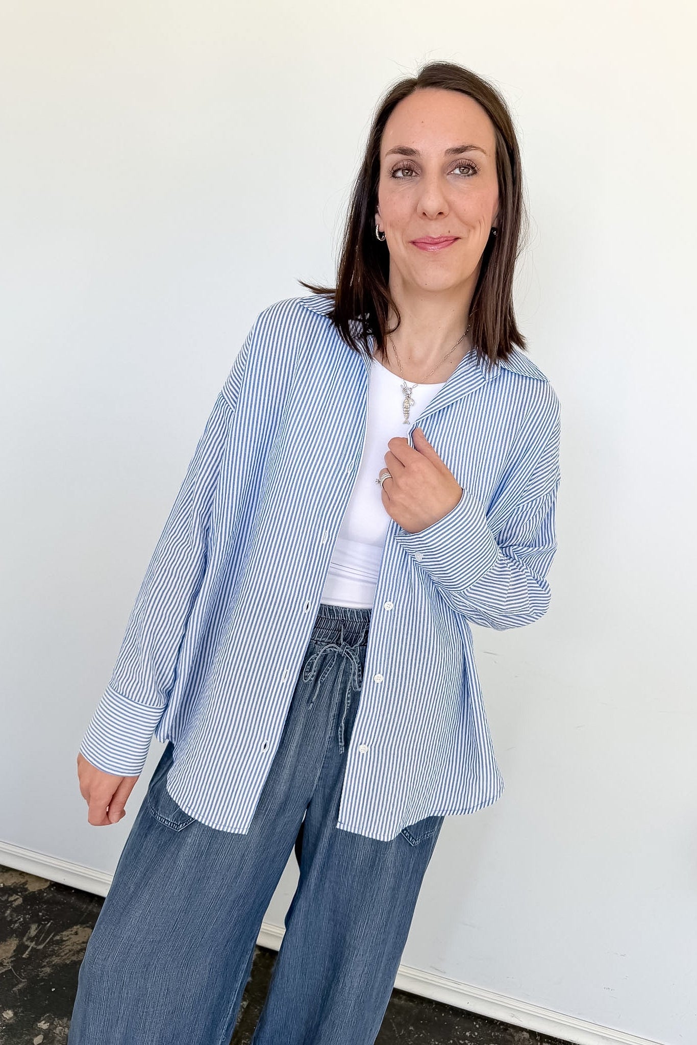 Woman wearing a blue striped shirt and gray pants against a white wall.