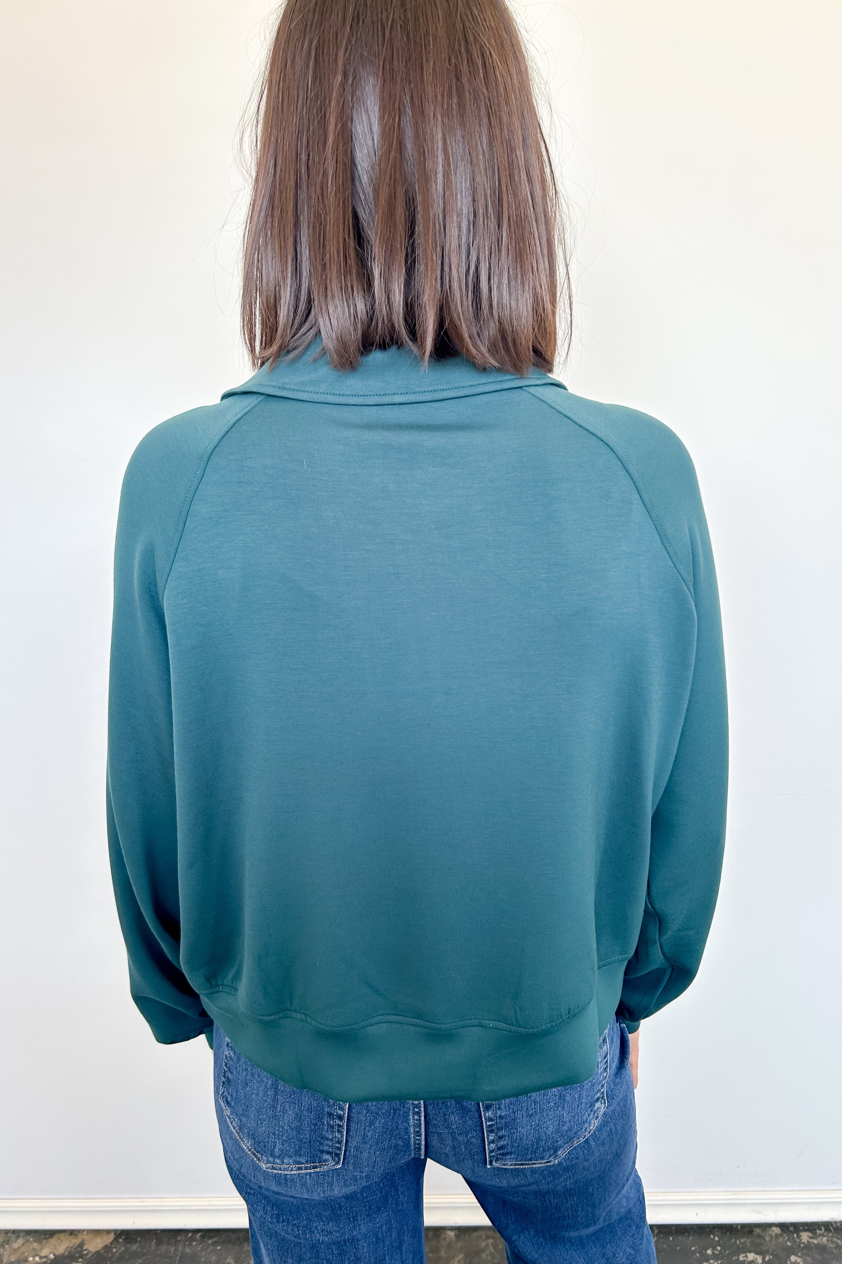 Person wearing a teal sweater and blue jeans against a white wall
