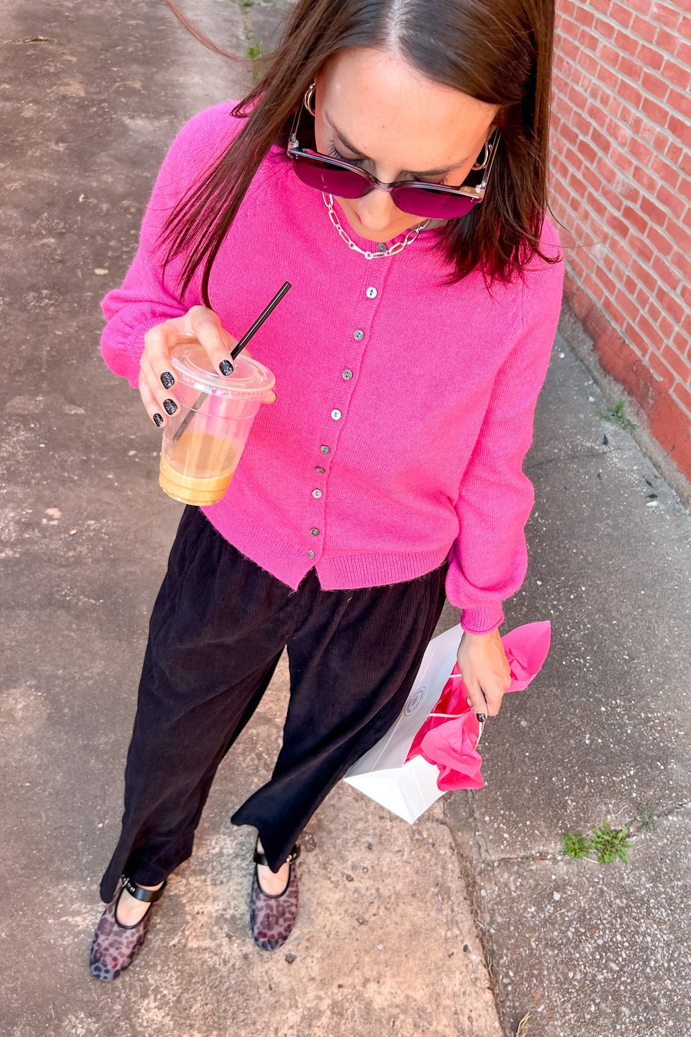 Woman in a pink cardigan and black pants holding a drink on a sidewalk.