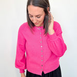Woman in hot pink cardigan standing up against a white wall.