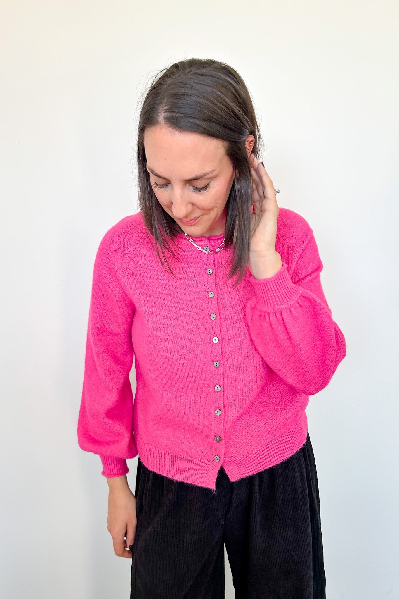 Woman in hot pink cardigan standing up against a white wall.