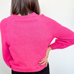 Woman in hot pink cardigan standing backward in front of a white wall with her hand on her hip.