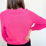 Woman in hot pink cardigan standing backward in front of a white wall with her hand on her hip.