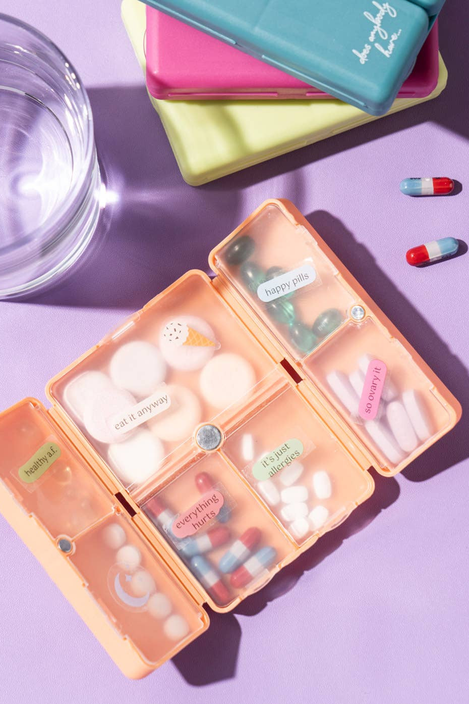 Colorful pill organizer with assorted pills and a glass of water on a purple background
