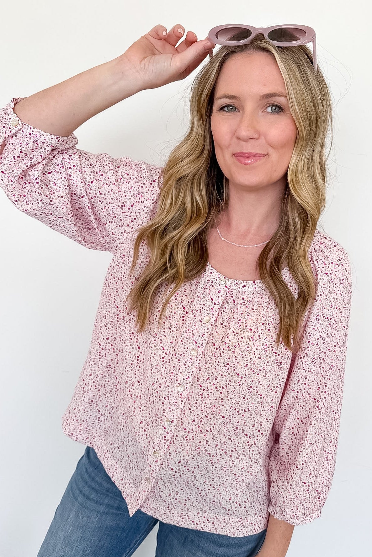 Woman wearing a pink floral blouse and blue jeans, holding sunglasses on a white background