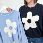 Woman holding a light blue sweater with a white flower design next to a black sweater with a similar design.