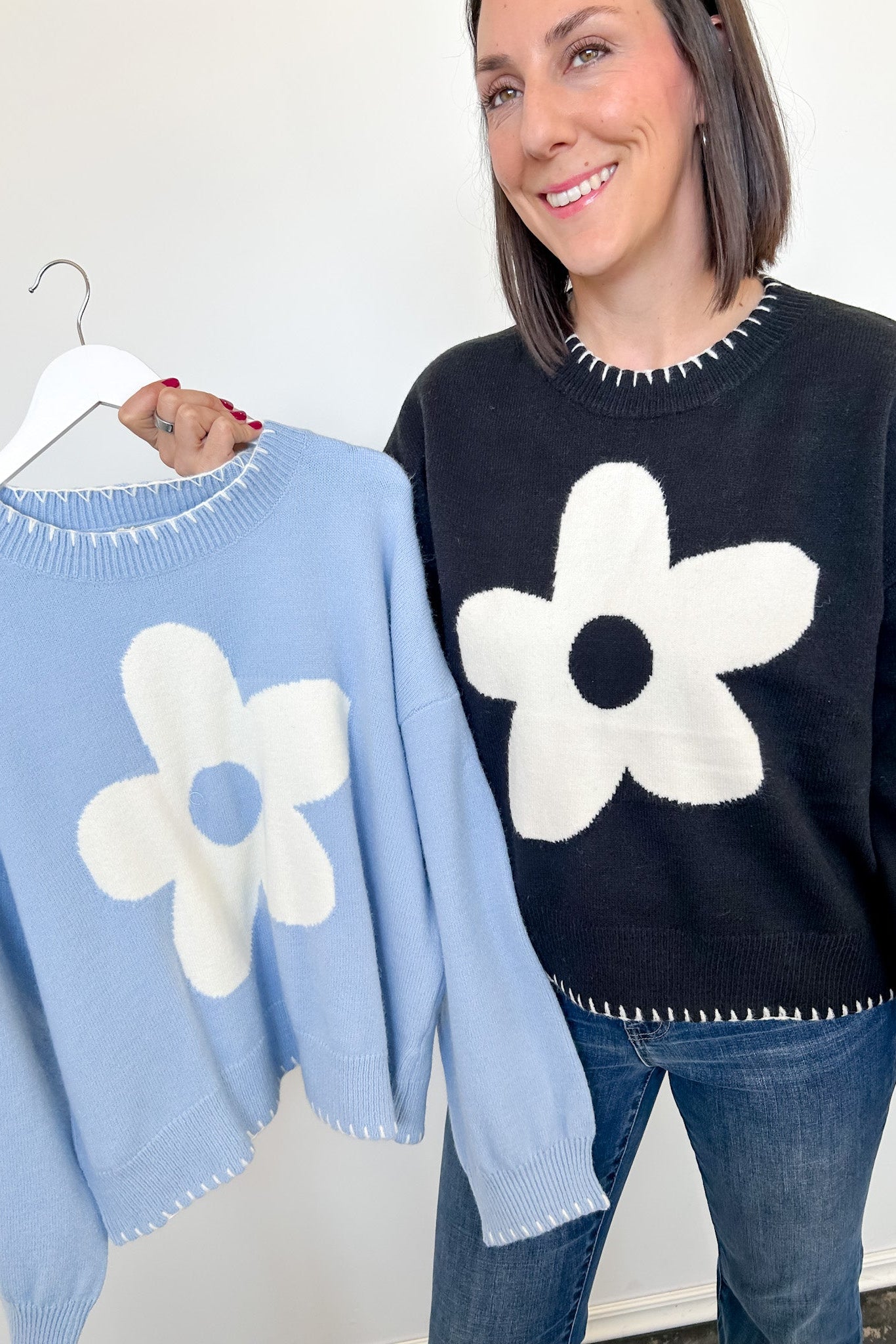 Woman holding a light blue sweater with a white flower design next to a black sweater with a similar design.