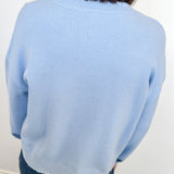 Person wearing a light blue sweater and jeans against a white wall.