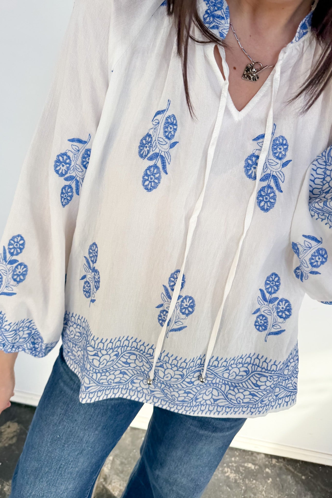White blouse with blue floral patterns worn by a person, standing against a neutral background.
