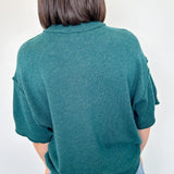 Person wearing a teal sweater and blue jeans against a white wall