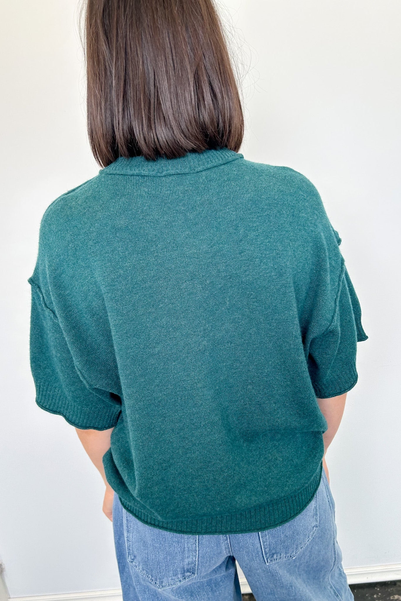 Person wearing a teal sweater and blue jeans against a white wall
