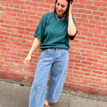 Person wearing a green shirt and blue jeans standing against a red brick wall.