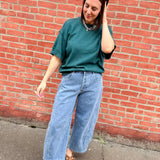 Person wearing a green shirt and blue jeans standing against a red brick wall.