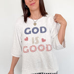Woman wearing a white t-shirt with 'GOD IS GOOD' text, standing against a white wall.