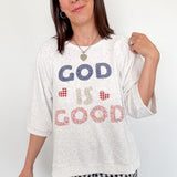 Woman wearing a white t-shirt with 'GOD IS GOOD' text, standing against a white wall.