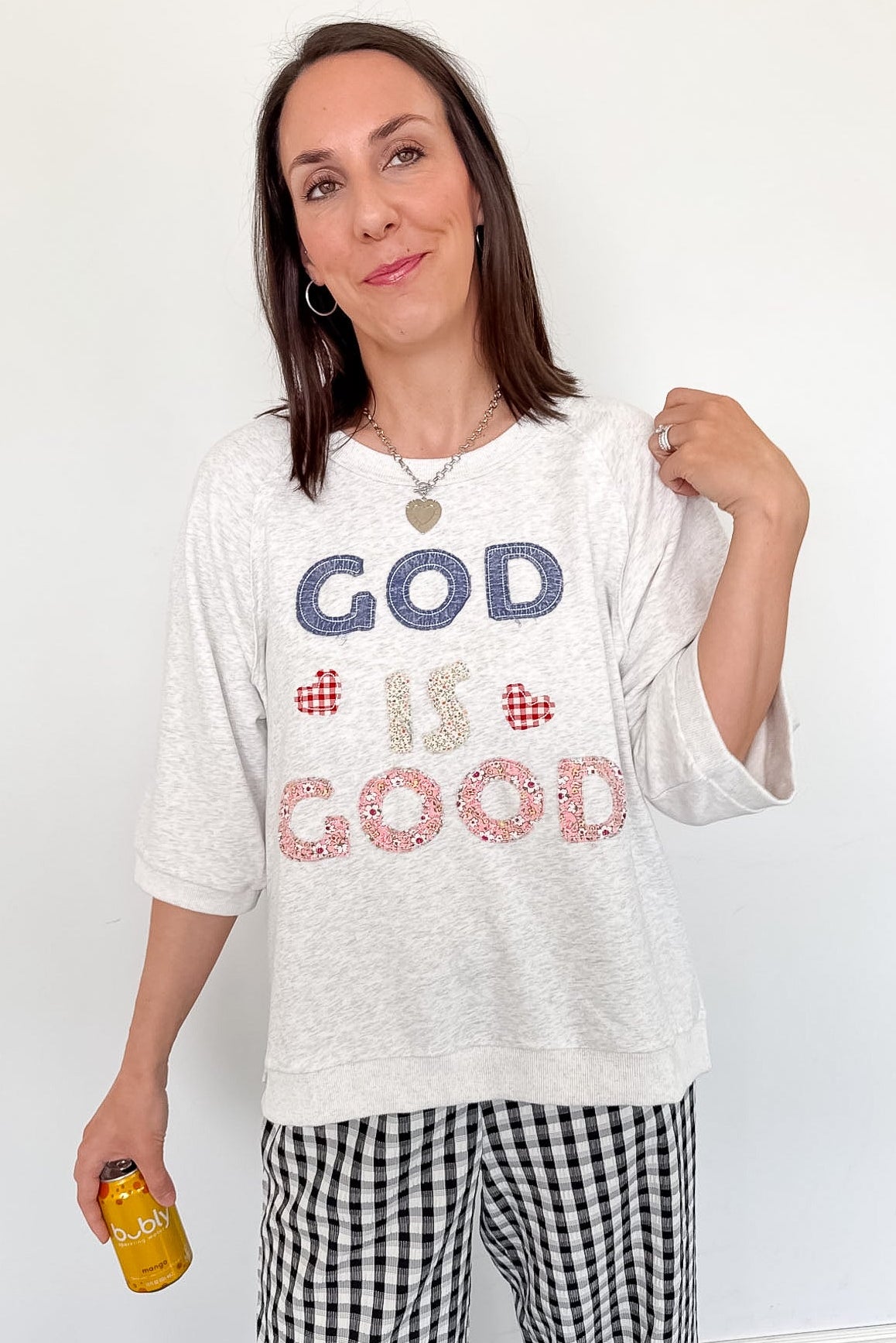 Woman wearing a white t-shirt with 'GOD IS GOOD' text, standing against a white wall.