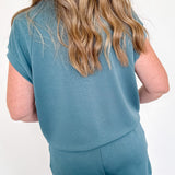 Person wearing a teal outfit with shorts against a white wall