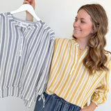 Woman holding a striped shirt on a hanger next to herself wearing a yellow and white striped shirt.